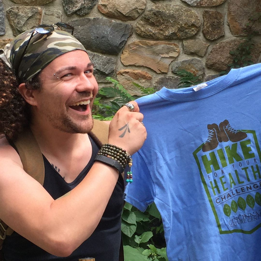 A young white man wearing a bandana around his head smiles widely as he holds up a Hike for Your Health t-shirt